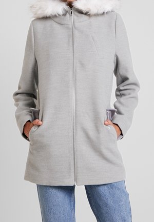 Manteau gris clair zippé avec bordure de capuche en fausse fourrure blanche, mains dans les poches, porté avec un jean bleu sur un fond neutre.