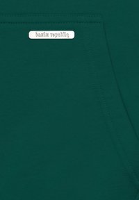 Sötétzöld szövet, sima textúrával, varrott zsebkészítéssel és egy ezüst címkével, amelyen az "bazix republiq" felirat áll. Nincsenek látható minták vagy díszítések.