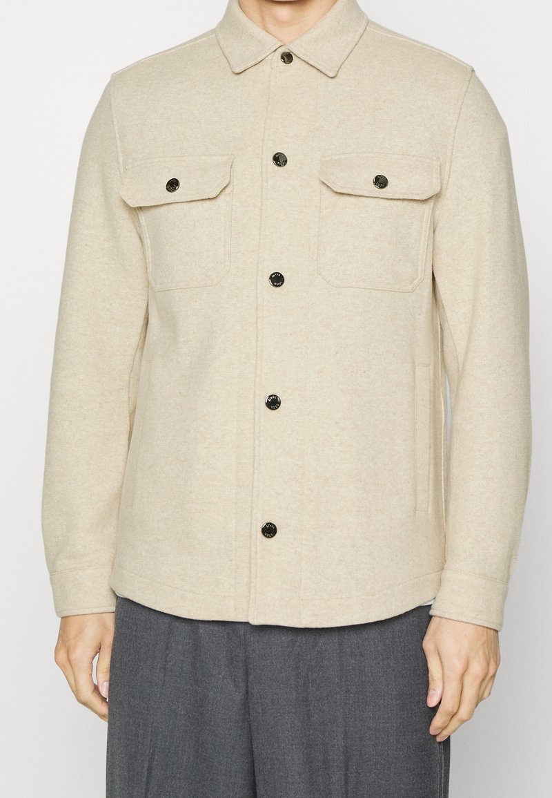 Beige wollen shirtjacket met knoopsluiting aan de voorkant, twee borstzakken en zwarte hardware-accenten; ontspannen pasvorm met een rechte zoom en lange mouwen.