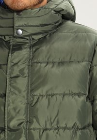 Grön vadderad jacka med glänsande yta, hög krage, tryckknappar och horisontell quiltad design. Inga fickor synliga.