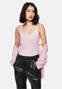 Helles rosa Tanktop mit einem runden Ausschnitt, kombiniert mit schwarzen Jogginghosen aus Kunstleder mit elastischem Bund. Weicher rosa Pullover darüber drapiert.