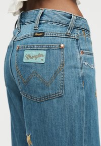 Κοντινό πλάνο μπλε τζιν Wrangler που φοριέται στους γοφούς, εμφανίζοντας την πίσω τσέπη με την ετικέτα Wrangler και τις χαλκιές καρφίτσες.