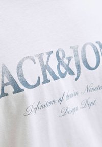 Λευκό βαμβακερό t-shirt με μπλε γραφιστικό κείμενο που αναγράφει "ACK & JON" και "Definition of denim. Design Dept." σε διαφορετικές γραμματοσειρές.