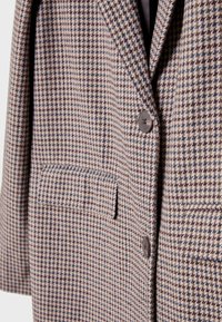 Blazer a motivi houndstooth marrone e beige con rever a punta, due bottoni e una tasca sul petto. Dettagli di tessuto testurizzato visibili.