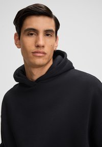 Felpa nera con tessuto texturizzato, dotata di un grande cappuccio, vestibilità comoda e design minimale. Esibita su uno sfondo neutro.
