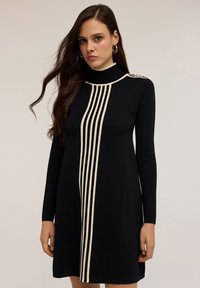 Abito nero con maniche lunghe, collo alto e strisce bianche verticali. Presenta dettagli decorativi sulle spalle e una texture liscia.