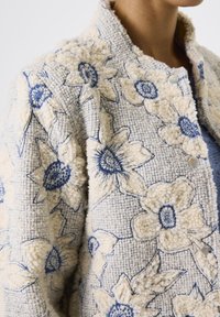 Cremfarbener, strukturierter Jacke mit blauen, gestickten Blumenmustern, Druckknöpfen und hohem Kragen, getragen von einer Person, die vom Hals abwärts zu sehen ist.