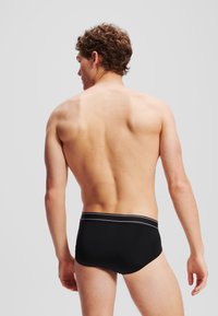 Slip de înot negru cu o curea contrastantă albă cu dungi, material neted și un design ajustat, evidențiind un aspect minimalist și aerodinamic.