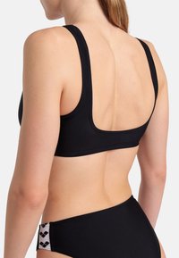 Svart bikiniöverdel med breda band och låg rygg. Materialet är slätt och elastiskt, med enkla linjer. Underdelen har en mönstrad detalj på sidan.