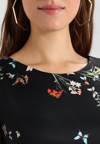 Femme portant de grandes boucles d'oreilles géométriques en or et un haut noir avec des motifs floraux colorés et des papillons près de l'encolure.