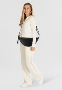 Borsa a tracolla nera in pelle, con design curvo, tracolla regolabile e dettaglio con nappina, indossata da un modello in abbigliamento chiaro e sneaker bianche.