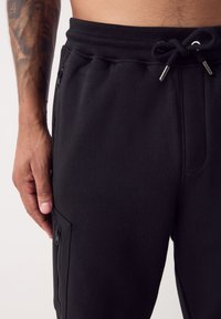 Zwarte joggerbroek gemaakt van zacht materiaal met een elastische tailleband, trekkoordsluiting en zijzakken met rits. Soepele textuur, aansluitend ontwerp.