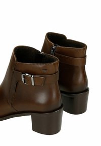 Stivali ankle in pelle marrone con punta arrotondata, chiusura con zip e un accento in fibbia metallica. Presentano un tacco a blocco e una fodera texturizzata.