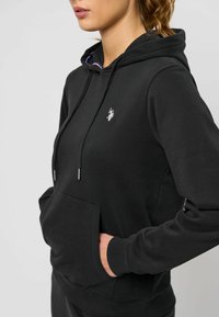 Zwarte hoodie met een voorzak, featuring een klein wit logo op de borst en een textuurstof. De trekkoorden hebben zilveren accenten.
