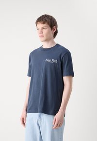Navyblauwe katoenen T-shirt met korte mouwen en een ronde hals. Beschikt over witte tekst "New York" op de bovenste linker borst.