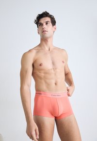 Bovenlijfs man in koraal kleurige Calvin Klein boxershorts, kijkt omhoog tegen een eenvoudige witte achtergrond.