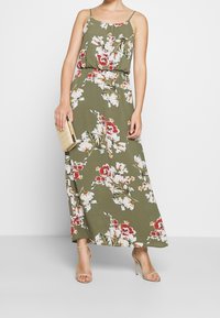 Maxi mekko oliivinvihreänä, jossa on valkoisia ja punaisia kukkakuoseja. Ominaisuuksiin kuuluu ohuet hihnat ja rento malli; yhdistetty kullanväriseen clutchiin ja korsettikorkokenkiin.