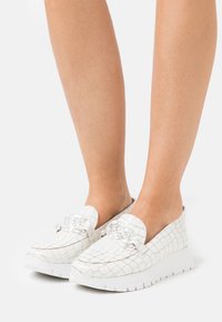 White slip-on sneakers with a crocodile texture, featuring a clear embellishment on the front and a chunky white sole.