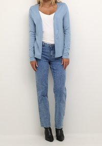 Donna che indossa un cardigan azzurro chiaro, una maglietta bianca a scollo a V, dei jeans a vita alta blu e stivali neri alla caviglia, in piedi contro un muro semplice.