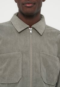 Nahaufnahme einer Person, die eine hellgraue, gerippte Cordjacke mit zwei Brusttaschen und einem silbernen Reißverschluss vorne trägt, Kragen sichtbar.