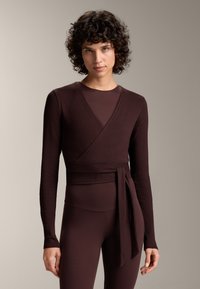 Tamno smeđa dugosleeve wrap majica s rebrastom teksturom, V-izrezom i vezanom strukom, u kombinaciji s odgovarajućim prilagođenim donjim dijelom.