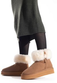 Botas de ante marrón hasta el tobillo con un puño esponjoso beige, punta redondeada y suela de goma plana, combinadas con medias negras y un vestido de punto verde oscuro.