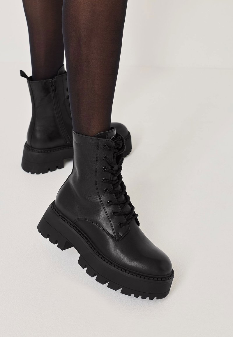 Bottes de cheville en cuir noir avec une semelle épaisse et robuste. Elles comprennent des lacets, une fermeture éclair sur le côté et une plateforme texturée pour une meilleure traction.
