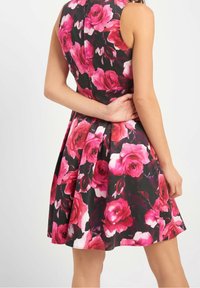 Černé šaty bez rukávů s živými vzory růžových a červených růží, s upnutým pasem a rozšířenou sukní, zobrazené ze zadní strany s jednou rukou za tělem.