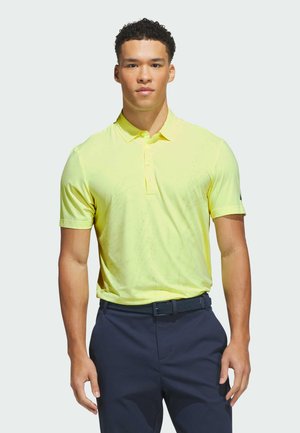Hombre con una camisa polo de manga corta de color amarillo claro metida en pantalones azul marino con un cinturón oscuro, de pie frente a un fondo liso.