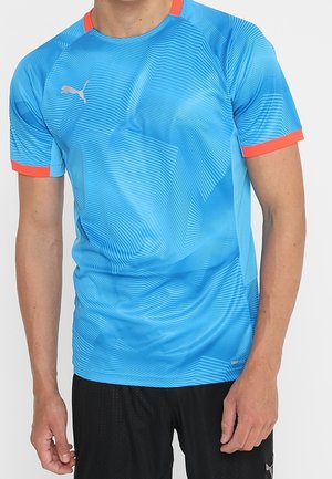 Hombre que lleva una camiseta deportiva azul de Puma con ribete rojo en las mangas y el cuello, combinada con pantalones cortos negros.