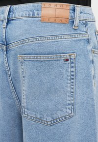 Ανοιχτό μπλε τζην παντελόνι με δερμάτινη ετικέτα πίσω που γράφει «TOMMY JEANS». Περιλαμβάνει πίσω τσέπη με μικρό λογότυπο σε κόκκινο, λευκό και μπλε χρώμα.