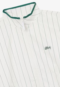 Biała kurtka na zamek błyskawiczny z cienkimi, ciemnymi pionowymi paskami, zielonym obszyciem na kołnierzu oraz małym haftowanym logo "Blkvis 13" na piersi.