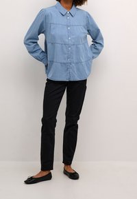 Ljusblå denimskjorta med knappar och krage, med vertikala sömmar och långa ärmar, i kombination med svarta slim-fit byxor och svarta lägenheter.