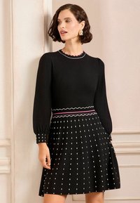 Vestito nero con lunghe maniche a sbuffo; parte superiore texturizzata con scollatura a zig-zag. La gonna plissettata presenta un motivo a pois bianchi e una vita a contrasto.