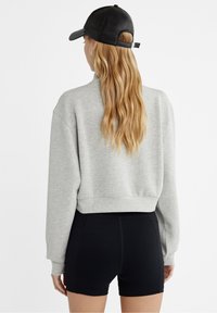 Kvinna med långt blont hår som bär en svart keps, grå avkortad sweatshirt och svarta tighta shorts, sedd bakifrån mot en vit bakgrund.