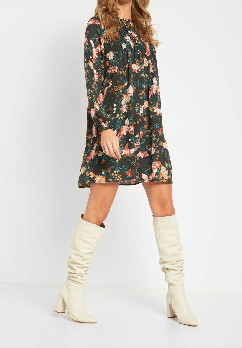 Robe à imprimé floral avec manches longues, encolure froncée et coupe ample. Caractérisée par une base vert foncé ornée de fleurs roses, orange et turquoise. Portée avec des bottes blanches montantes jusqu'aux genoux.