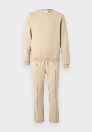 Bluză bej și pantaloni de jogging asortați, confecționați din material moale. Bluza are gât rotund și manșete cu refile; pantalonii de jogging au cusături laterale.