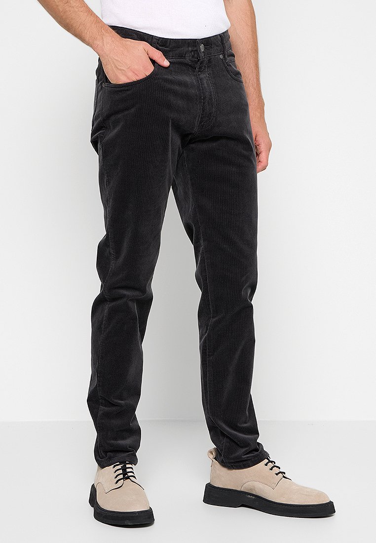 Mann trägt schwarze Cordhosen, beiger Schnürschuhe und ein weißes ärmelloses Shirt, steht mit einer Hand in der Tasche.