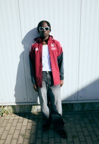 Röd och svart zip-jacka med logotyp, vit t-shirt, grå vida jeans, svarta skor och vita solglasögon. Skugga på en texturerad yta.