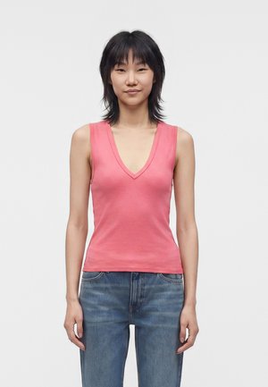 Jeune personne aux cheveux courts et foncés portant un haut rose sans manches à col en V et un jean bleu, debout devant un fond blanc uni.
