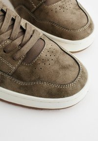 Brązowe zamszowe sneakersy z perforowanym czubkiem, teksturowanymi brązowymi akcentami z tkaniny, białą śródpodeszwą i kontrastowym detalem przeszycia.