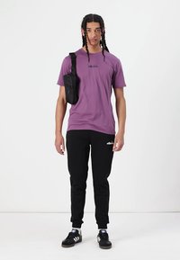 Fialové bavlnené tričko s logom, spojené s čiernymi teplákmi a čiernymi teniskami. Crossbody taška visí z ľavého ramena. Jednoduchý, ležérny štýl.
