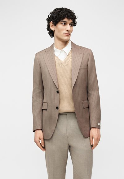 Άνδρας με μπεζ σακάκι με ρίγες, πάνω από μπεζ πλεκτό γιλέκο και λευκό πουκάμισο με γιακά, και ανοιχτό γκρι παντελόνι, στραμμένος ελαφρώς προς τα αριστερά.