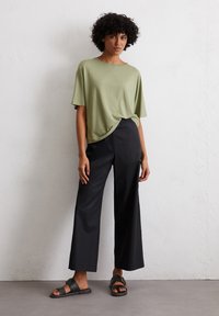 Olijfgroene top met korte mouwen, een losse pasvorm en knoopdetail aan de onderkant, gecombineerd met zwarte wijde broek en zwarte sandalen.