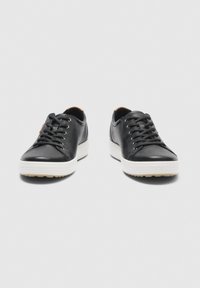 Par de zapatillas bajas de cuero negro con suelas blancas gruesas y cordones negros, vistas desde el frente sobre un fondo gris.