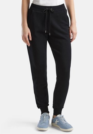 Mujer con pantalones jogger negros con cintura ajustable y zapatillas azul claro, de pie contra un fondo liso.