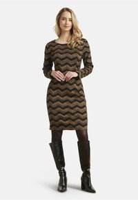 Langdurige jurk met een zigzagpatroon in zwart, goud en bruin. Gemaakt van een gestructureerde stof, tot de knie, gecombineerd met hoge zwarte laarzen.