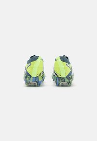 Scarpe da calcio con una tomaia gialla neon, dettagli in rete testurizzata e una suola a fantasia blu vivace. Presentano un tallone sagomato e tacchetti per grip.