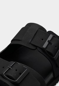 Μαύρο σανδάλι με δύο ρυθμιζόμενους ιμάντες με μεταλλικές αγκράφες, ματ υφή και ανοιχτή μύτη, που παρουσιάζει έναν μινιμαλιστικό σχεδιασμό.