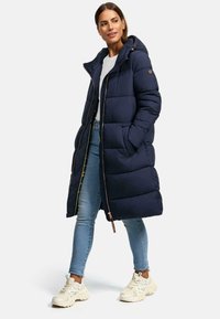 Navy puffer coat with a hood, featuring a zip closure, quilted design, and khaki lining. Paired with light blue jeans and cream sneakers.